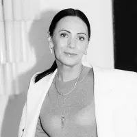 Woman with dark hair in a ponytail, wearing a light blazer and layered necklaces, smiles softly. The black-and-white image conveys a serene, elegant tone.