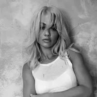 A woman with long, wavy hair leans against a textured wall. She wears a white sleeveless top. The black-and-white image conveys a calm, introspective mood.