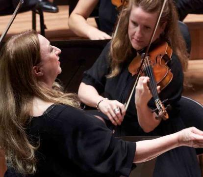 A conductor leads an orchestra, gesturing with a baton, while musicians around her play violins.