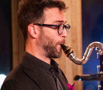 A man wearing glasses and a dark suit plays a saxophone.