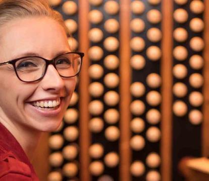 A smiling person wearing glasses sits at an organ, with music sheets.