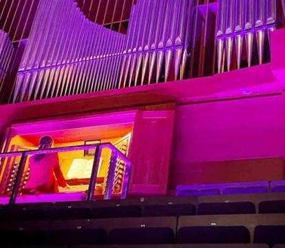 A large pipe organ illuminated by purple and pink lights, with a person seated at the organ console.