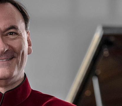 Stephen Hough wears a red shirt standing in front of a grand piano