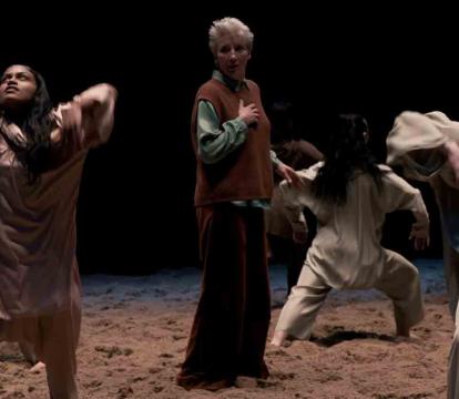 A group of people in loose, earth-toned clothing dance expressively on a sandy stage.