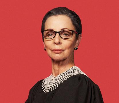 A white woman in her 60s has her hair sleekly pulled back and pursed lips wears black glasses and a lawyer's black robe with white crocheted neck piece. She stands in front of an all-red background.