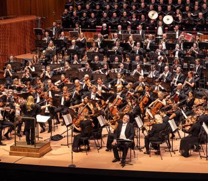 Simone Young stands with arms to the side in front of the Sydney Symphony Orchestra with a full choir insider the Concert Hall