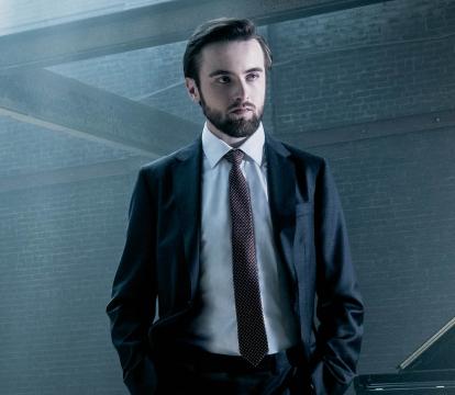 Pianist Daniil Trifonov stands in a suit in front of a grand piano