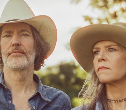 A man holding a guitar and woman both in cowboy hats look pensive away from the camera exuding a country music vibe.
