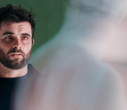 A white man with black hair and dark beard wears all black. He's in front of a green background and is looking at a shadow.