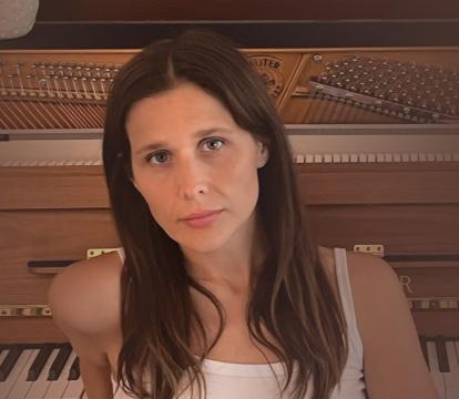 A woman in a white tank top and jeans leans against an upright piano with exposed strings. A white vase sits atop the piano.