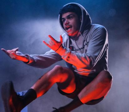 A boy in a hoodie and shorts performs a high jump on stage, arms expressive. Blue and red lights create a dynamic and dramatic atmosphere.