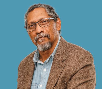 A black man stares at the camera, he wears black glasses and has a beard and grey hair.