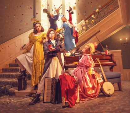 Five women stand staggered on a staircase in regency era gowns, with musical instruments for Pride and Prejudice (sort of) as confetti falls around them.