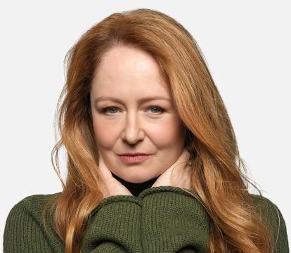 A woman with long red hair wearing a green sweater, holding her hands up under her face against a white background.