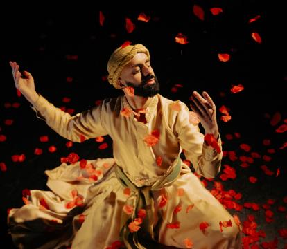 A man in traditional Persian attire gracefully dances amidst swirling red rose petals against a dark background.