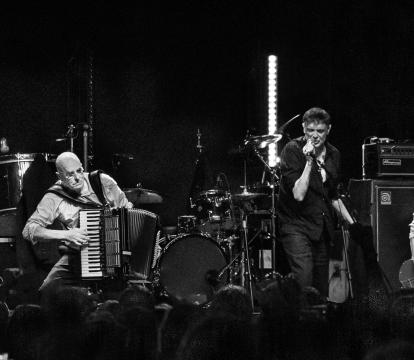 Black and white photo of a lively folk band performing on stage. Musicians play diverse instruments, including guitar, accordion, and banjo.