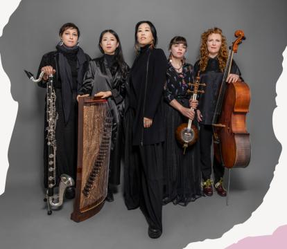 Five female musicians in black attire stand confidently against a gray backdrop. They hold various instruments, including a bass clarinet, global string instruments, and cello.