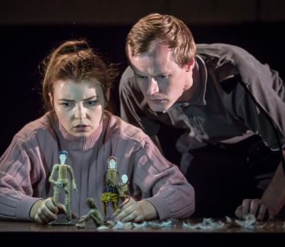 A man and a woman stare intently at small puppets of people.