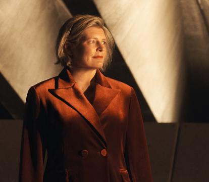 A woman wearing a dark orange jacket looking off to the side, with concrete beams in the background.