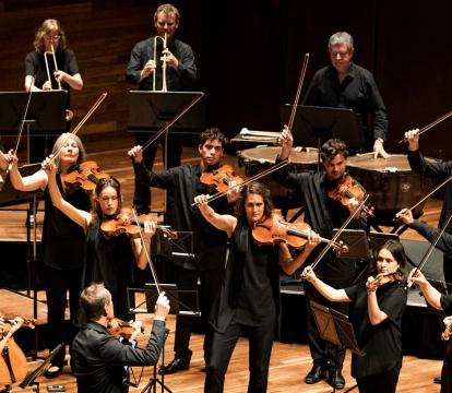 The string section of an orchestra on stage mid-performance, all wearing black, with bows raised.