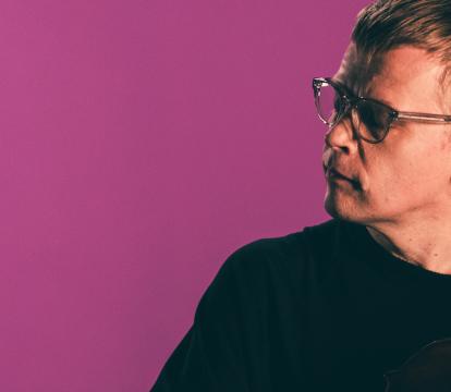 A man wearing a black t-shirt and glasses holding a violin against a magenta coloured background.