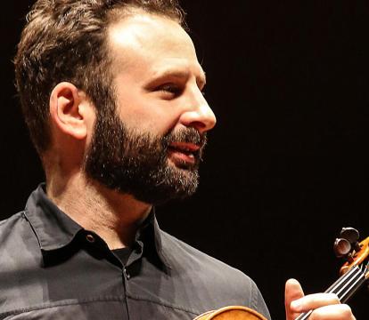 A cropped portrait of a man holding a violin against a dark backdrop.