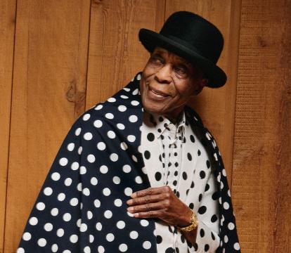 A 90-year-old African American man wears a bowler hat and black and white spotty outfit.