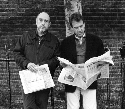 Two men with newspapers, standing in front of a brick wall and iron gates.