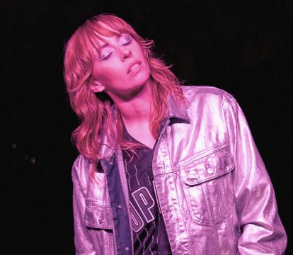 A white woman in her 30s wears a pink reflective jacket and meditatively closes her eyes. She's bathed in pink light.