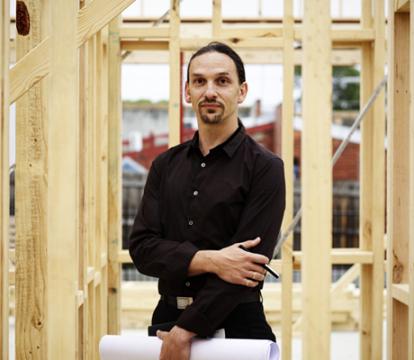 A man in a black shirt and long hair standing underneath wooden construction work.