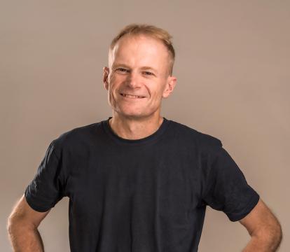 A man with short light brown hair smiles with his hands on his hips. He wears a black t-shirt.