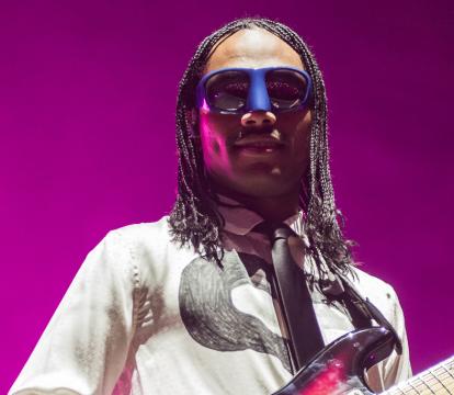 A person wearing googles and a white long sleeve t-shirt playing a guitar against a bright purple background.