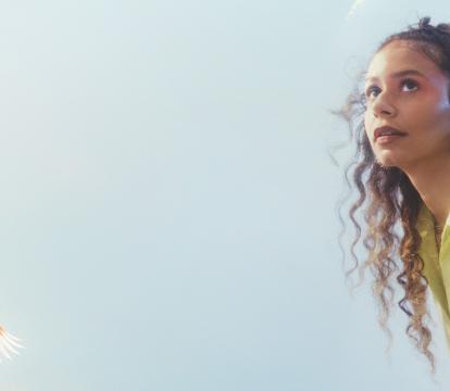A performer wears a light green blouse with pink eyeshadow looks off camera, the moon and a bird is seen behind her