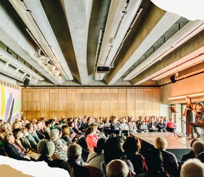 A group of people seated in the Utzon Room watching a performer on stage.