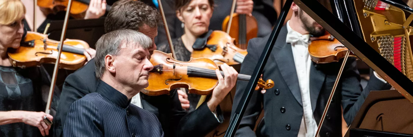 Stephen Hough wears a pinstripe shirt as his eyes are closed playing the piano in front of the violin section of an orchestra