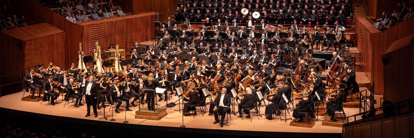 Simone Young stands with arms to the side in front of the Sydney Symphony Orchestra with a full choir insider the Concert Hall