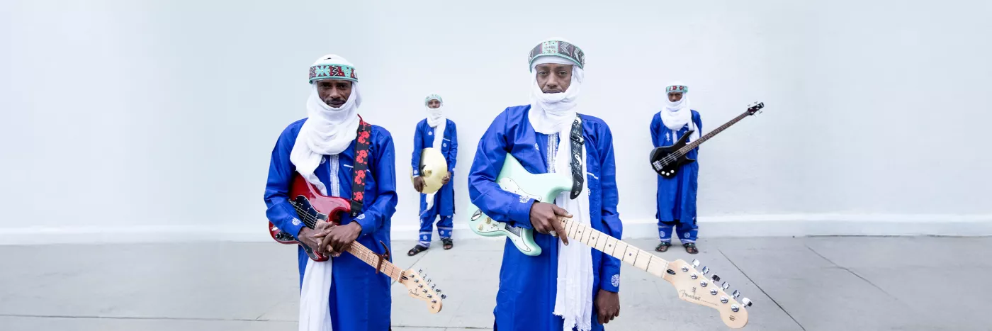 Four men wearing blue outfits and white headscarves, three holding a guitar and one holding a drum cymbal.