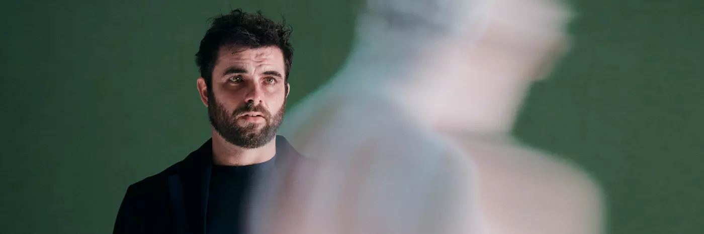 A white man with black hair and dark beard wears all black. He's in front of a green background and is looking at a shadow.