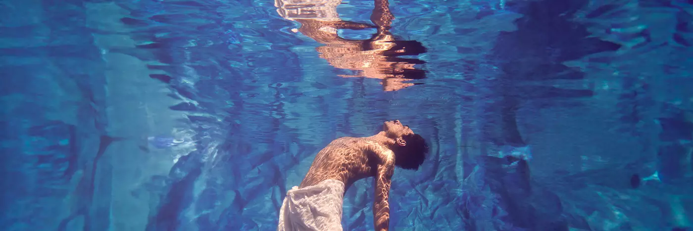 A shirtless man swimming underwater in a pool wearing a cloth skirt has his face looking up towards the sky,