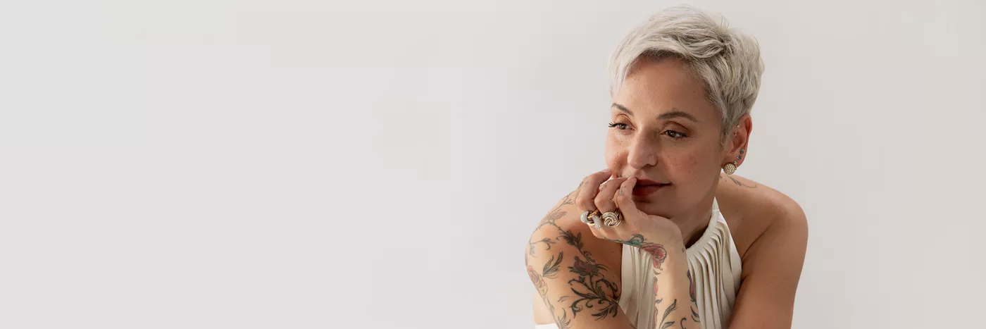 A woman adorned with tattoos with short, bleached blonde hair is comfortably positioned on a chair, resting her chin in her hand.