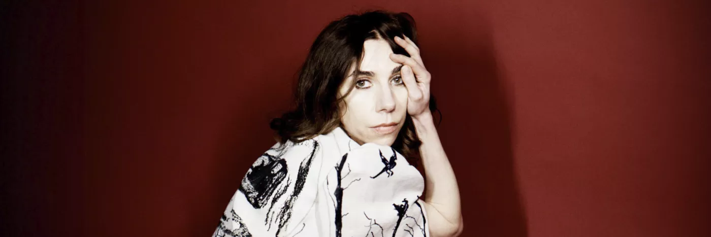 A white woman with long brown hair wears a stylish white dress with a black splash pattern. She's crouched down and looking at the camera.