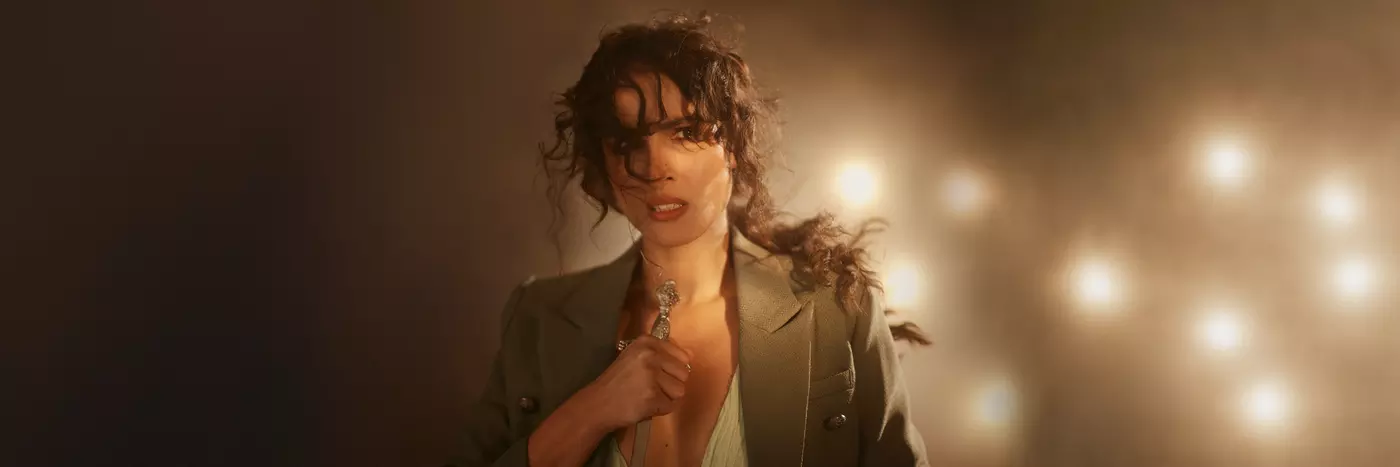 A white woman with a messy, curly ponytail and wearing a beige blazer looks worried at the camera while holding the handle of a sword.