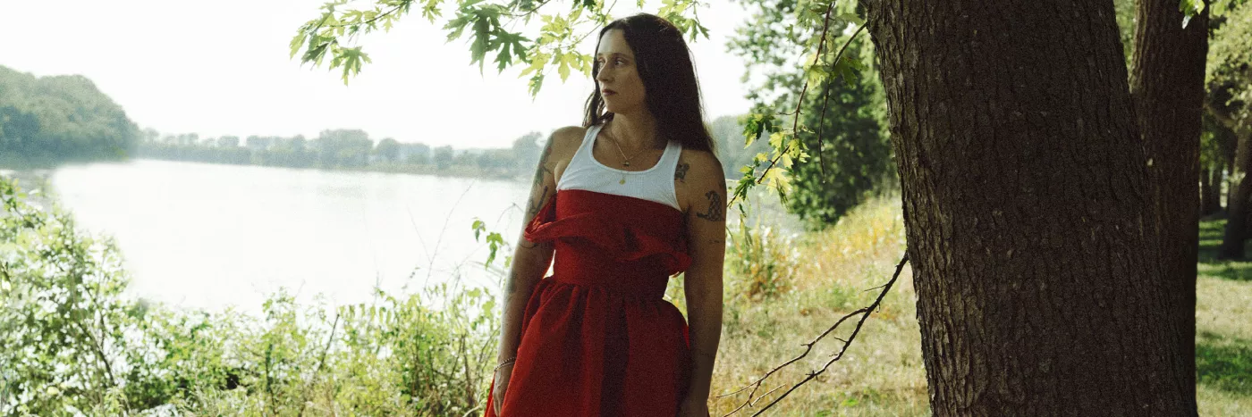 Woman in red dress standing by river, gazing into distance.