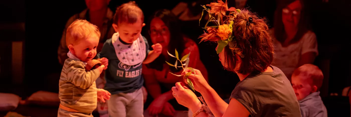 A women wearing a crown of flowers is holding up some leaves to two toddlers.