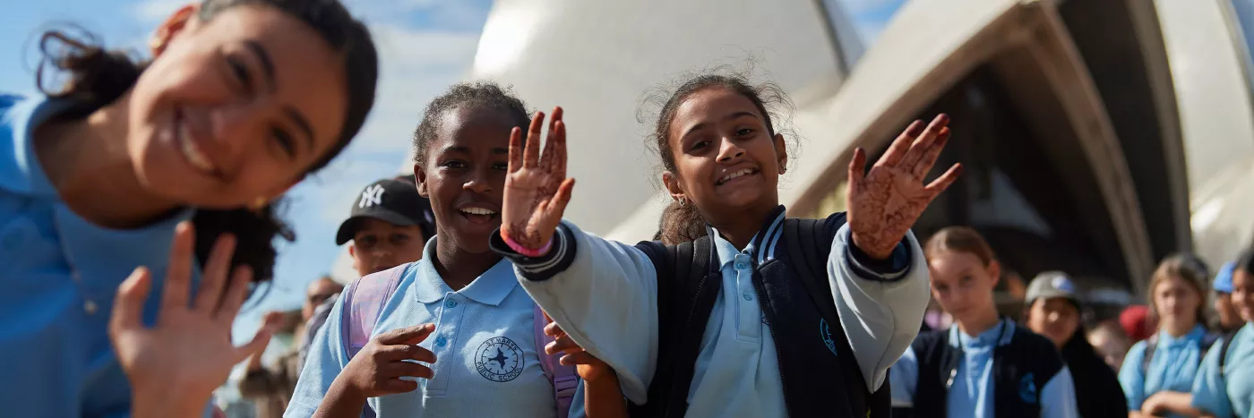 A group of girls wave at the camera in school uniform. The Sydney Opera House sails are seen blurry in the background.