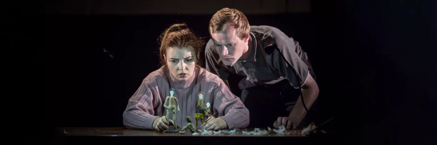 A man and a woman stare intently at small puppets of people.