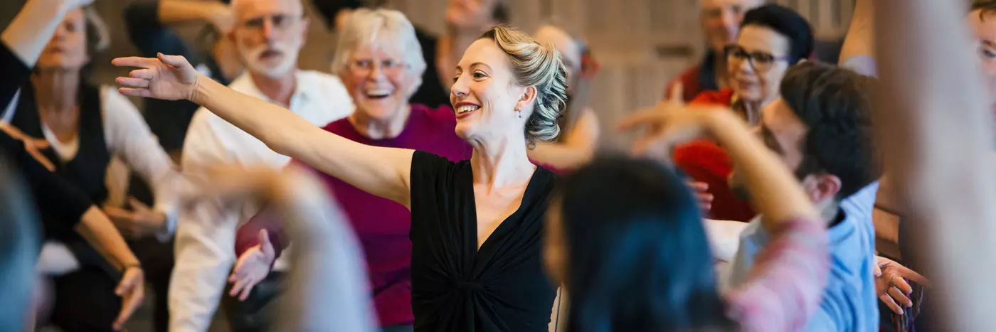 A woman in a black dress smiling, reaching out with an audience surrounding her.