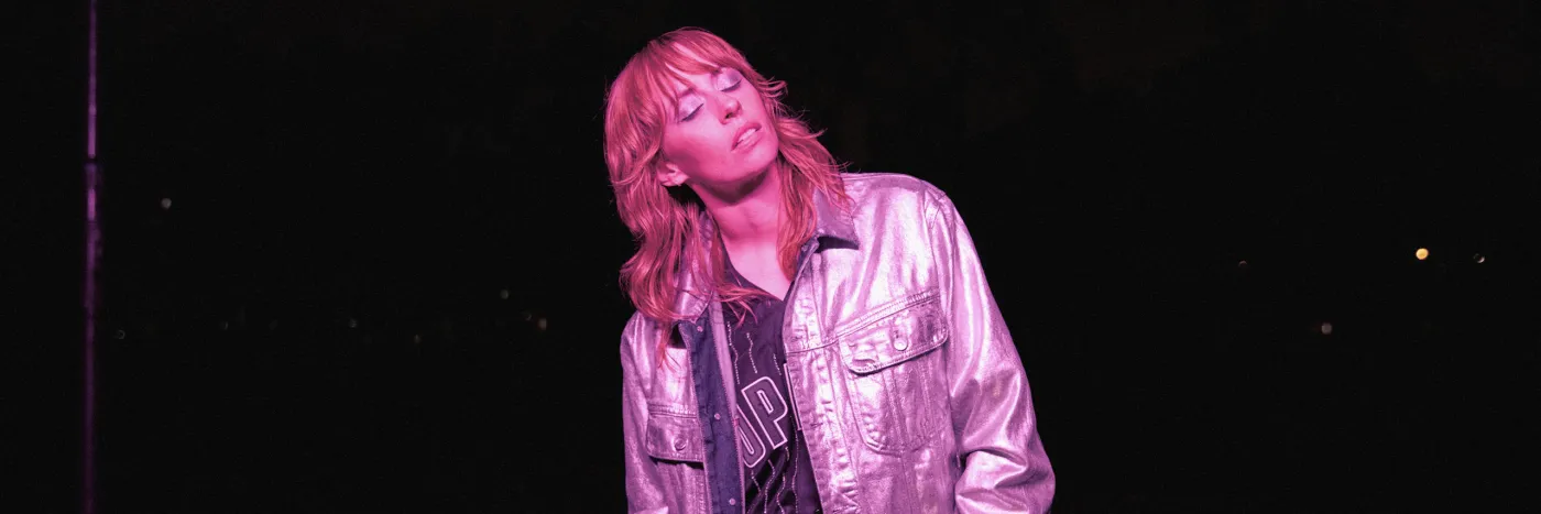 A white woman in her 30s wears a pink reflective jacket and meditatively closes her eyes. She's bathed in pink light.