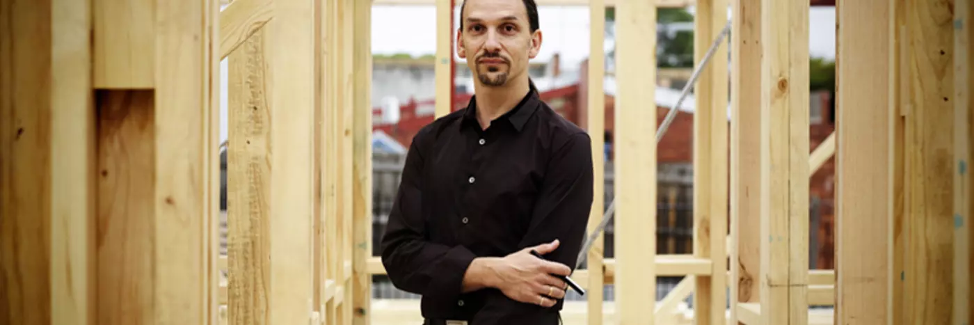 A man in a black shirt and long hair standing underneath wooden construction work.