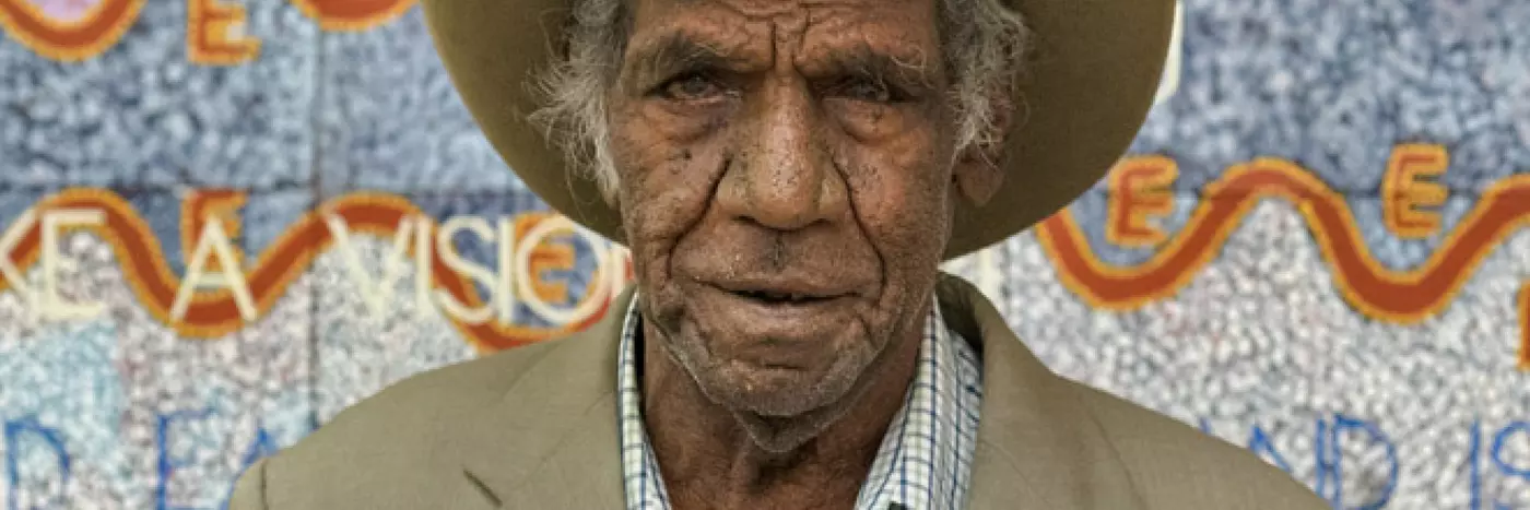 Michael Nelson Jagamara wearing a beige suit jacket over a blue and white checked shirt and wide brim hat, standing in front of an artwork in pastel colours.